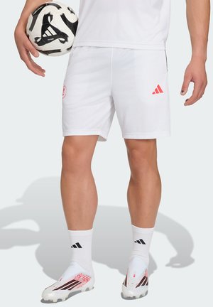 Idrettsutøver iført hvite shorts, sokker og fotballsko som holder en svart-hvitt Adidas fotball ved siden av seg.