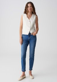 Gilet sans manches en lin blanc avec un col en V et fermeture par bouton, associé à un jean en denim bleu et des ballerines crème. Design simple et ajusté.