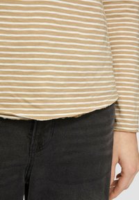 Gestreiftes Langarmoberteil in Hellbraun und Weiß, mit abgerundetem Saum und strukturiertem Stoff, kombiniert mit schwarzen Jeans.