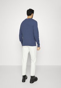 Marineblaue gerippte Strickjacke mit Rundhalsausschnitt, im Rücken mit einem roten und weißen Akzent versehen. Getragen mit weißen Hosen und schwarzen Stiefeletten.