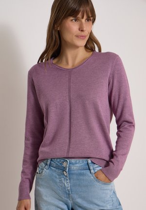 Mujer con un suéter morado de manga larga y vaqueros azul claro, con la mano derecha en el bolsillo, mirando ligeramente hacia un lado.