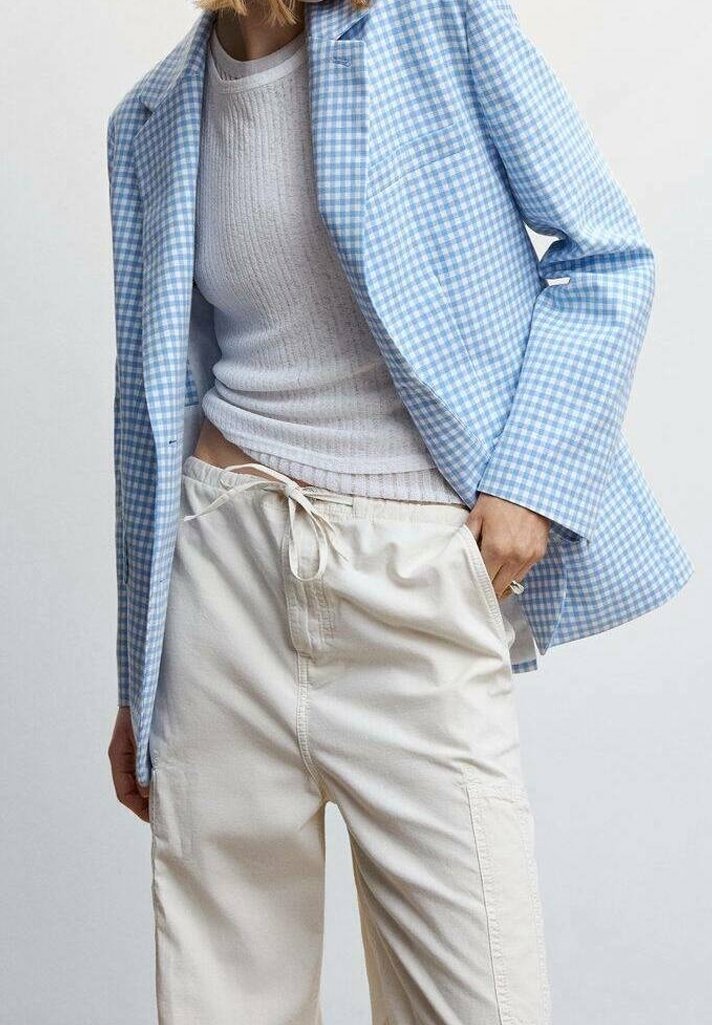 Personne portant un blazer vichy bleu clair, un top blanc à manches longues côtelé et un pantalon crème à cordon, main dans la poche sur fond uni.