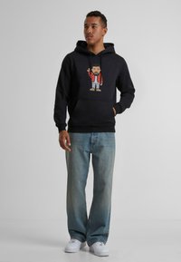 Zwarte hoodie met een cartoonafbeelding aan de voorkant, met een figuur in een rode jas. Gekozen in combinatie met lichtblauwe brede jeans en witte sneakers.