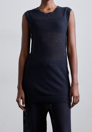 Bezrękawnikowa czarna dzianinowa tunika z okrągłym dekoltem, noszona na dopasowanych dzianinowych spodniach, ukazująca fakturowany, półprzezroczysty materiał.