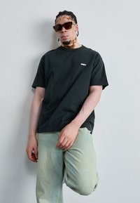Black cotton t-shirt with the white text "OBEY" on the chest, paired with light green denim pants featuring embroidered words "MOST" and "ALIVE."