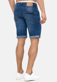 Shorts en jean bleu foncé avec un revers, incluant deux poches à l'arrière et une étiquette en cuir au niveau de la taille. Baskets blanches aux pieds.