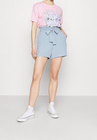Personne portant un short taille haute bleu clair avec une ceinture nouée, un t-shirt graphique Metallica rose et des baskets montantes blanches, se tenant avec les mains dans les poches.