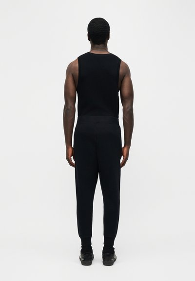 Άνδρας που στέκεται με την πλάτη προς τα εμάς, φοράει μαύρο αμάνικο τοπ, μαύρο παντελόνι και μαύρα αθλητικά παπούτσια, με απλό φωτεινό γκρι φόντο.