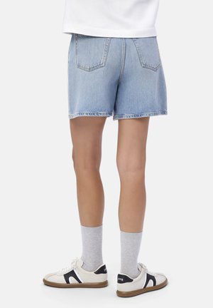 Person, der hellblaue Jeansshorts, graue Crew-Socken und weiße Sneakers mit schwarzen Akzenten trägt, steht vor einem einfarbigen Hintergrund.