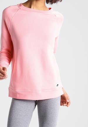 Vrouw draagt een lichtroze longsleeve sweater en grijze legging, staande tegen een effen lichtgrijze achtergrond.
