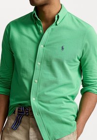 Grön pikétröja med långa ärmar, knäppkrage och broderad logotyp på bröstet. Tillverkad av strukturerad bomullstyg.