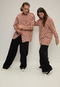 Dos personas llevan camisetas extragrandes con un patrón geométrico en marrón y blanco. Combinan las camisetas con pantalones de pierna ancha negros y zapatillas casuales.