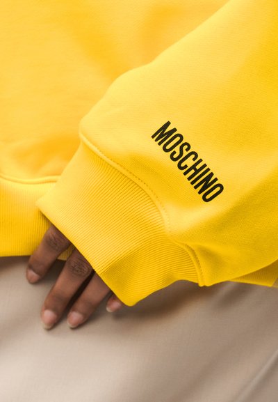 Main partiellement visible sous une manche de sweat-shirt jaune avec le texte noir "MOSCHINO" sur le poignet, reposant sur une surface en tissu clair.