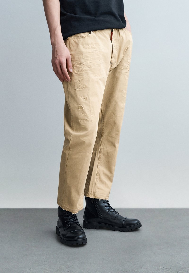 Jeune personne portant un pantalon beige usé avec des ourlets effilochés et des bottes de combat noires à lacets, debout sur un sol gris.