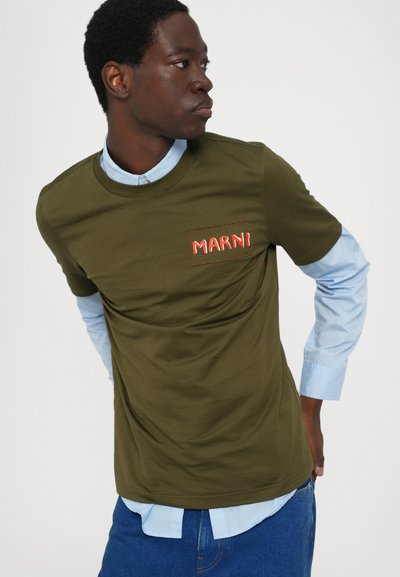 Tricou verde măsliniu cu guler rotund, având un logo "MARNI" cusut în roșu pe buzunarul stâng. Purtat peste o cămașă cu guler de culoare bleu deschis.