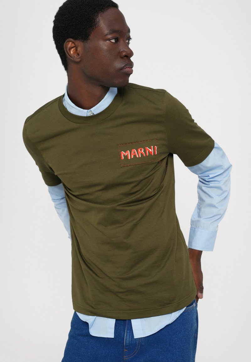 Camiseta verde oliva con cuello redondo, que presenta un logo bordado en rojo "MARNI" en el bolsillo izquierdo del pecho. Lleva una camisa de cuello azul claro debajo.
