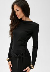 Czarna sukienka z długim rękawem z gładkiego materiału, o dopasowanej sylwetce, z drapowanym detalem po boku i złotymi akcentami na mankietach.