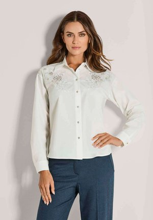 Witte langlopende blouse met knoopsluiting en borduursel op de schouders. Glad stof met een kraag en zilveren knoopaccenten.