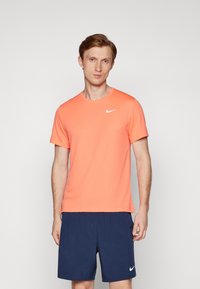 Nike kortærmet t-shirt i lys orange, lavet af letvægtsmateriale, med rund hals og et hvidt logo på venstre bryst. Sammen med navy shorts.