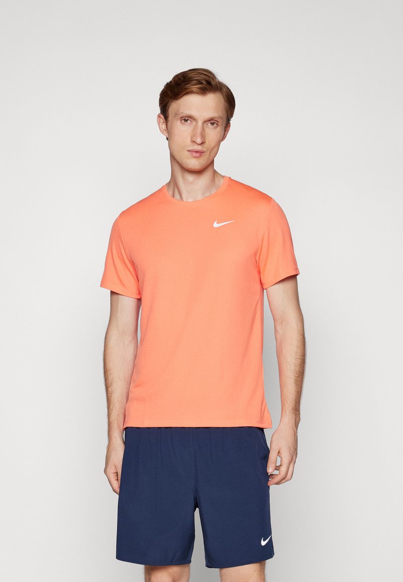 Nike kortærmet t-shirt i lys orange, lavet af letvægtsmateriale, med rund hals og et hvidt logo på venstre bryst. Sammen med navy shorts.