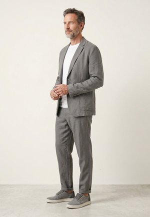 Mężczyzna stojący w szarym garniturze, białej koszuli i szarych trampkach na jednolitym jasnym tle.