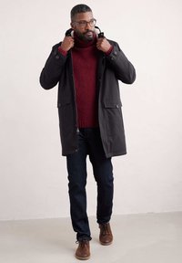 Parka noire avec une capuche, des poches avant et une fermeture éclair. Portée sur un pull bordeaux, associée à un jean bleu foncé et des chaussures brunes.