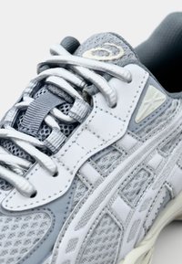 Zapatilla deportiva gris que presenta una parte superior de malla y cuero, con acentos blancos, cordones en contraste y una suela texturizada para mayor tracción.