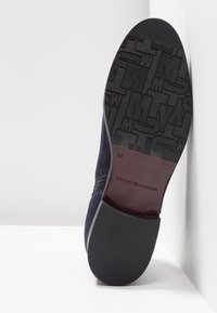 Semelle d'une chaussure en daim bleu foncé montrant une bande de roulement en caoutchouc à motif avec la marque « TOMMY HILFIGER » et le numéro de taille 37 embossé sur la section médiane marron.