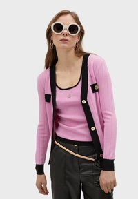 Rosa ribbad cardigan med svarta detaljer, med guldknappar och en ringad topp under, tillsammans med läderbyxor och överdimensionerade vita solglasögon.