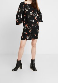 Žena v černých květinových šatech s volnými rukávy a černých kožených kotníkových botách stojící na světle šedém pozadí.