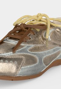 Gros plan d'une basket métallique argentée et bronze montrant deux lacets de couleurs différentes, un jaune et un marron, noués.