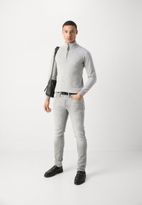 Pull à zip gris clair, jean gris clair fuselé et chaussures noires à enfiler. Le modèle tient un sac messager noir, debout contre un fond blanc.