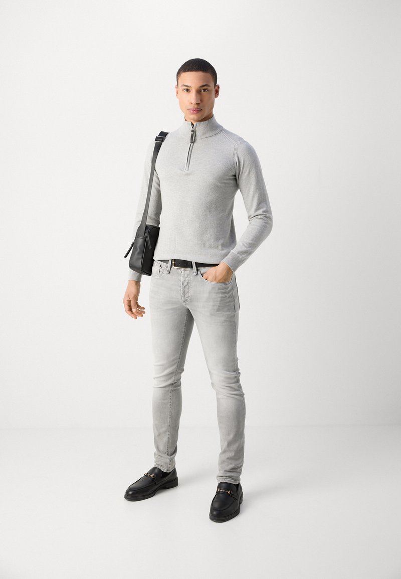 Maglione zip grigio chiaro, jeans grigio chiaro tapered e scarpe slip-on nere. Il modello tiene una borsa a tracolla nera, in piedi contro uno sfondo bianco.