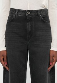 Gros plan d'une personne portant un jean en denim noir et un haut léger à manches longues, les mains détendues le long du corps, avec une peau foncée et du vernis à ongles rose.