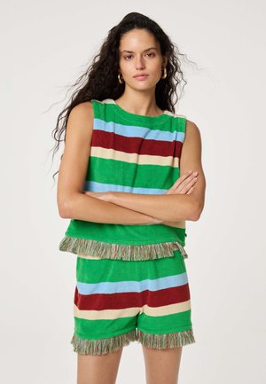Vrouw met lang krullend haar, draagt een mouwloos gestreept groen, rood, blauw en beige top en bijpassende korte broek met franjes, armen gekruist, neutrale achtergrond.