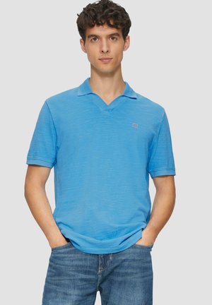 Camisa polo de manga corta azul con cuello en V, fabricada en tejido texturizado y con un pequeño logotipo en el pecho. Combinada con unos vaqueros azules.