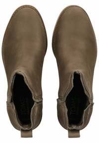 Botas de tobillo de cuero marrón con una textura suave, paneles laterales elásticos y detalles de costura. La marca TOMS es visible en el interior.
