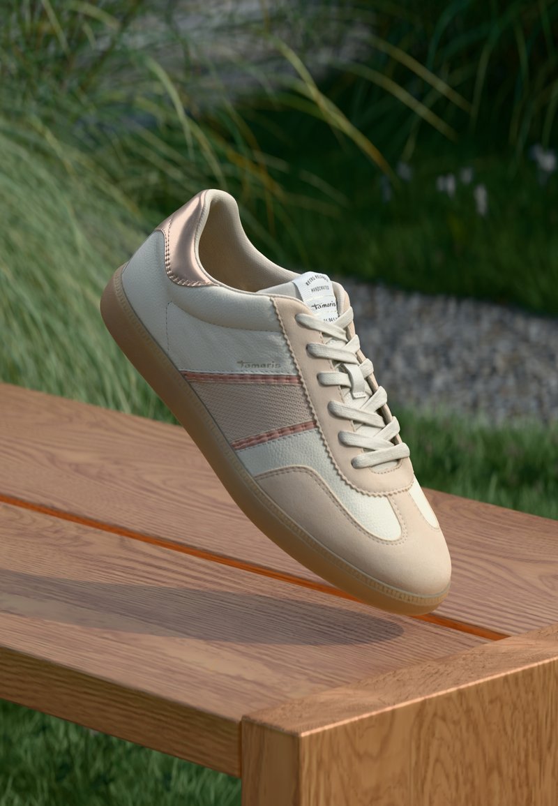 Baskets beige avec une tige en cuir lisse, accents rose clair et semelle en caoutchouc brun clair. Présente des lacets blancs et des rayures latérales discrètes.