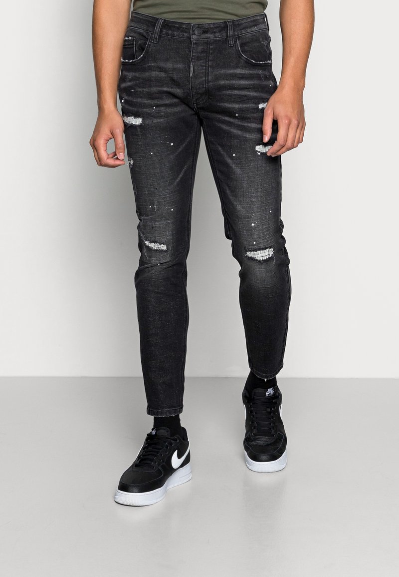 Jeans in denim nero con dettagli usurati, caratterizzati da strappi e sottili schizzi di vernice. Abbinati a sneaker nere e a una camicia verde scuro.