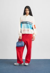 Bílý pulovr s vícebarevnými pruhy a logem "adidas", v kombinaci s červenými tepláky, šedými botami a modrou kabelkou s logem.