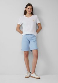 Witte T-shirt met korte mouwen met gele banaan borduursel, gecombineerd met lichtblauwe opgerolde shorts en witte sneakers met beige accenten.