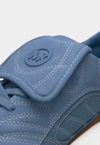 Μπλε αθλητικό παπούτσι με λουρί βέλκρο, λευκή ραφή, πλευρικό πάνελ από δίχτυ και στρογγυλό λογότυπο ανάγλυφο στο λουρί.