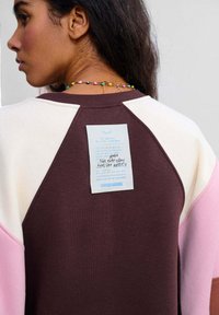 Didelio dydžio džemperis su bordo spalvos korpusu, kreminio ir rožinio raglano rankovėmis. Ant nugaros yra etiketė su ranka rašytu tekstu. Struktūruota medžiaga.