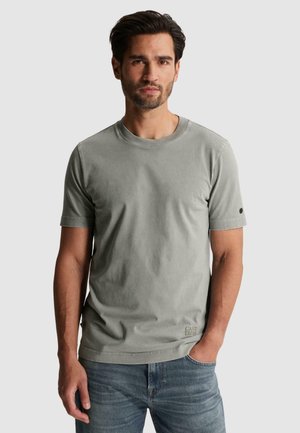 Mand med mørkt hår og skæg iført en enkel grå kortærmet t-shirt og blå jeans, stående med den ene hånd i lommen.