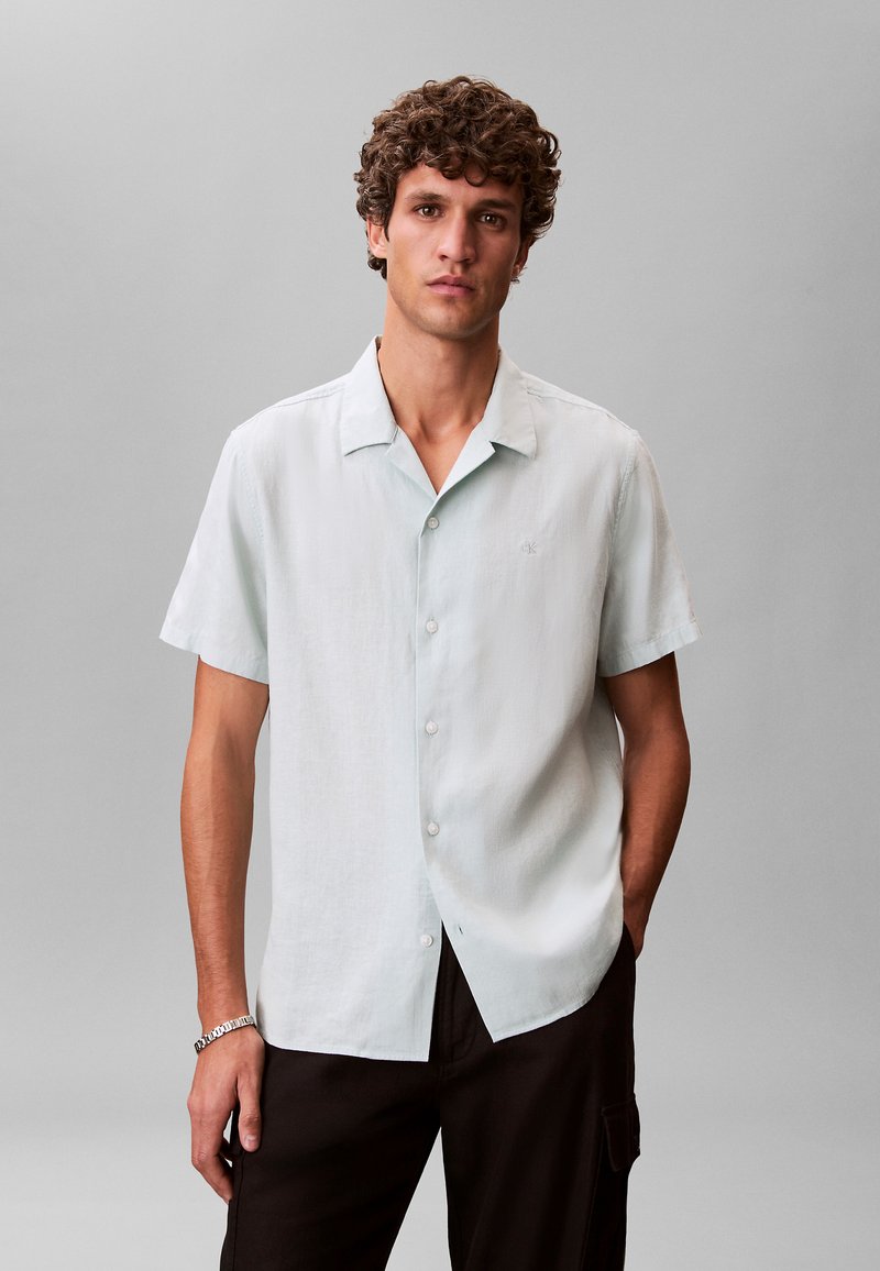 Joven con cabello rizado que lleva una camisa de manga corta azul claro abotonada y pantalones oscuros, de pie frente a un fondo gris liso.