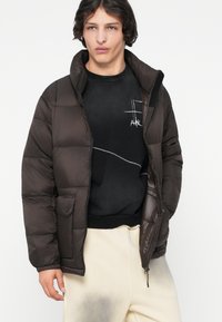 Barna puffertömlő, magas gallérral, cipzárral, elasztikus mandzsettákkal és egy pattentált zsebböl. Fekete grafikás pulóver és világos nadrág alatt viselve.