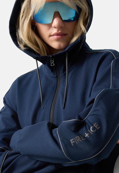 Person med blondt hår iført en navy hoodie med teksten "FIRE + ICE" på ærmet og reflekterende blå solbriller, krydsede arme mod en neutral baggrund.