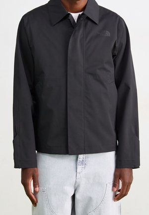 Homme portant une veste noire à fermeture éclair avec col et poches latérales, associée à un pantalon en jean bleu clair.