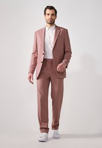 Hellrosa maßgeschneiderter Anzug mit einknopfiger Jacke, flachen Hosen und weißem Hemd, kombiniert mit weißen Sneakers.