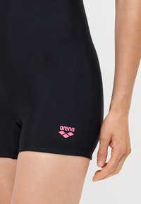 Maillot de bain noir au design épuré, fabriqué en matériau extensible. Il présente un logo "arena" rose sur la jambe gauche, sans motifs ni accents.